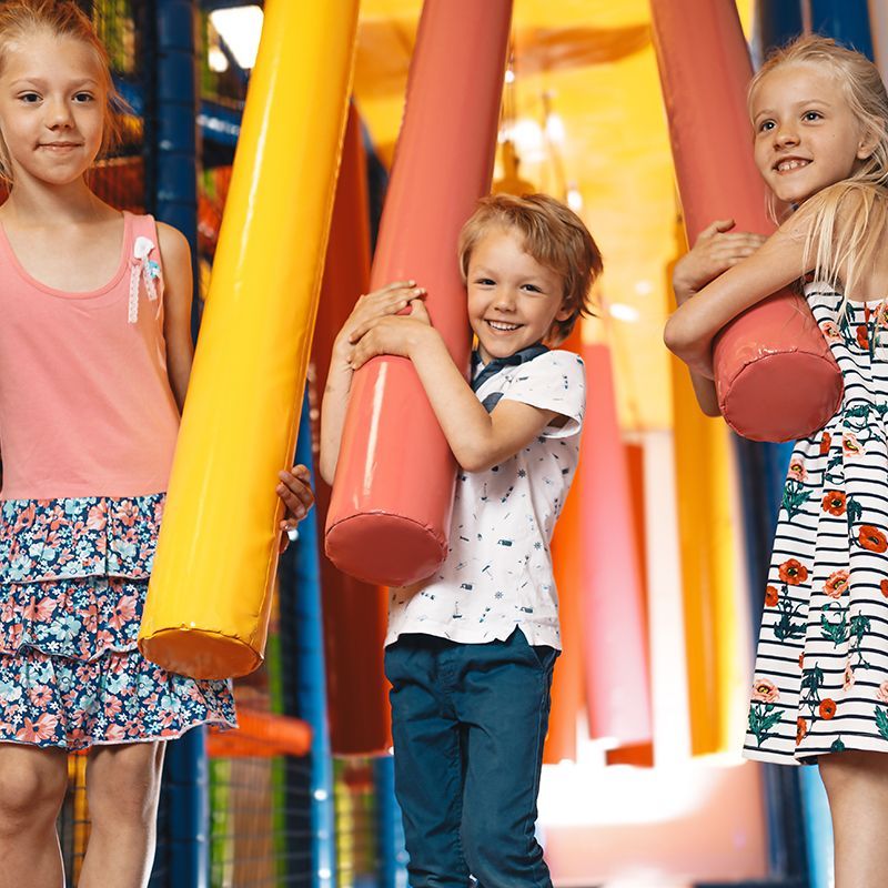 Three children smiling, holding large, colorful, padded pillars in a play area.