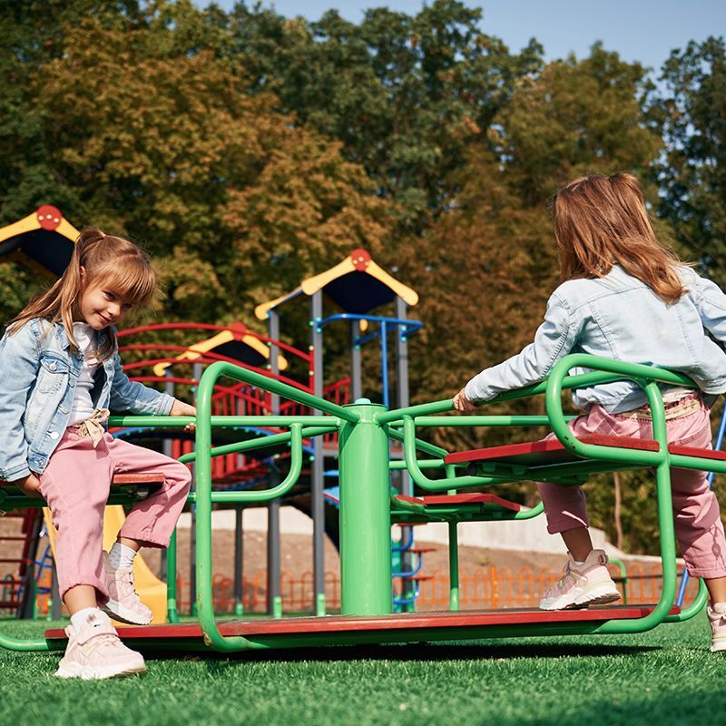 Two girls playing on a green and red merry-go-round at a playground.