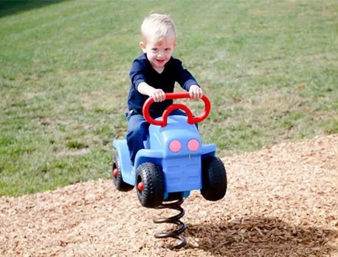 Boy riding a blue spring toy car on wood chips, smiling in a park.
