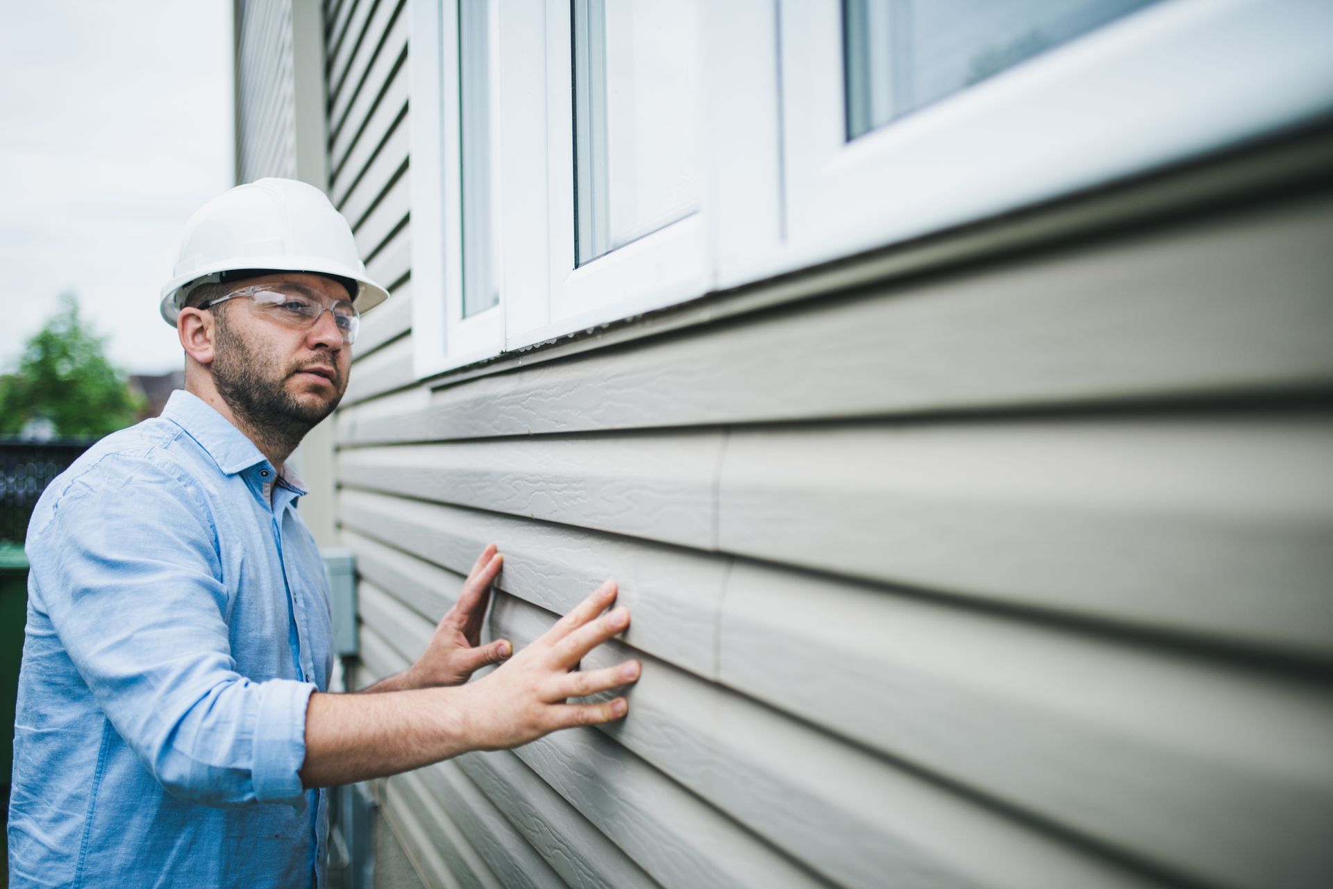 A man wearing a hard hat and safety glasses is inspecting the siding of a house.