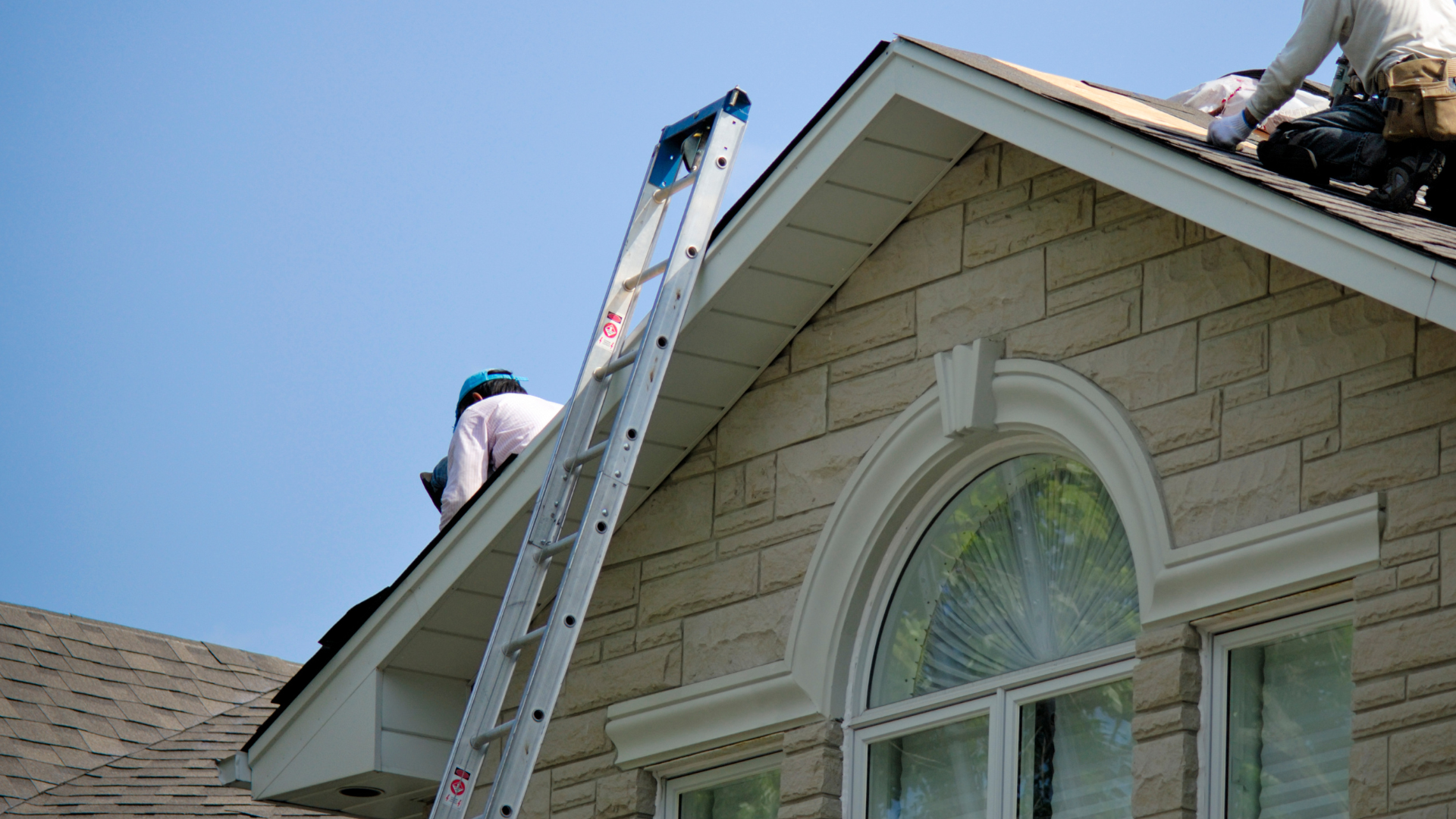 Workers on a roof, using a ladder. House has beige siding and a large arched window.
