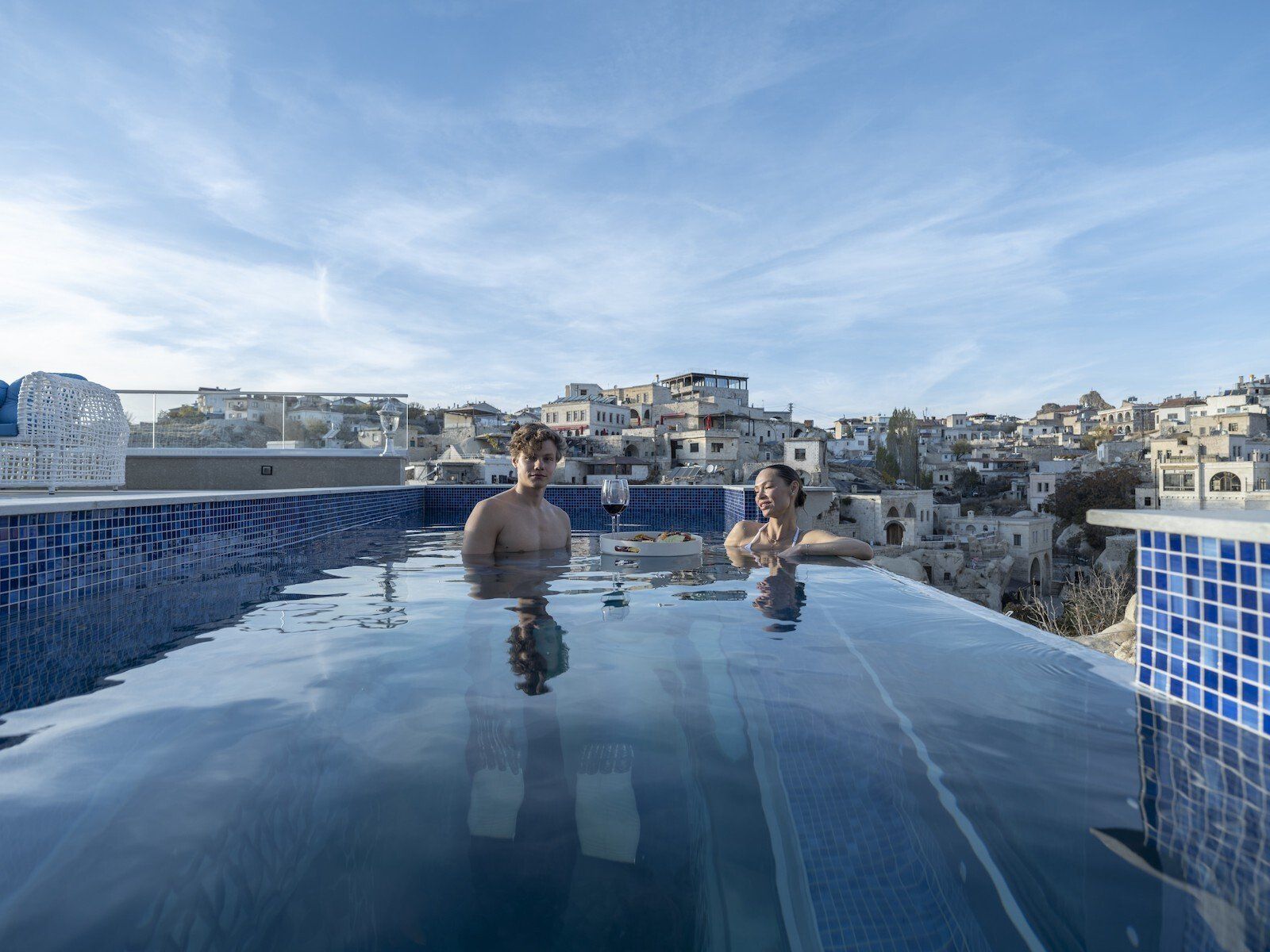 Two men are swimming in an infinity pool with a city in the background.