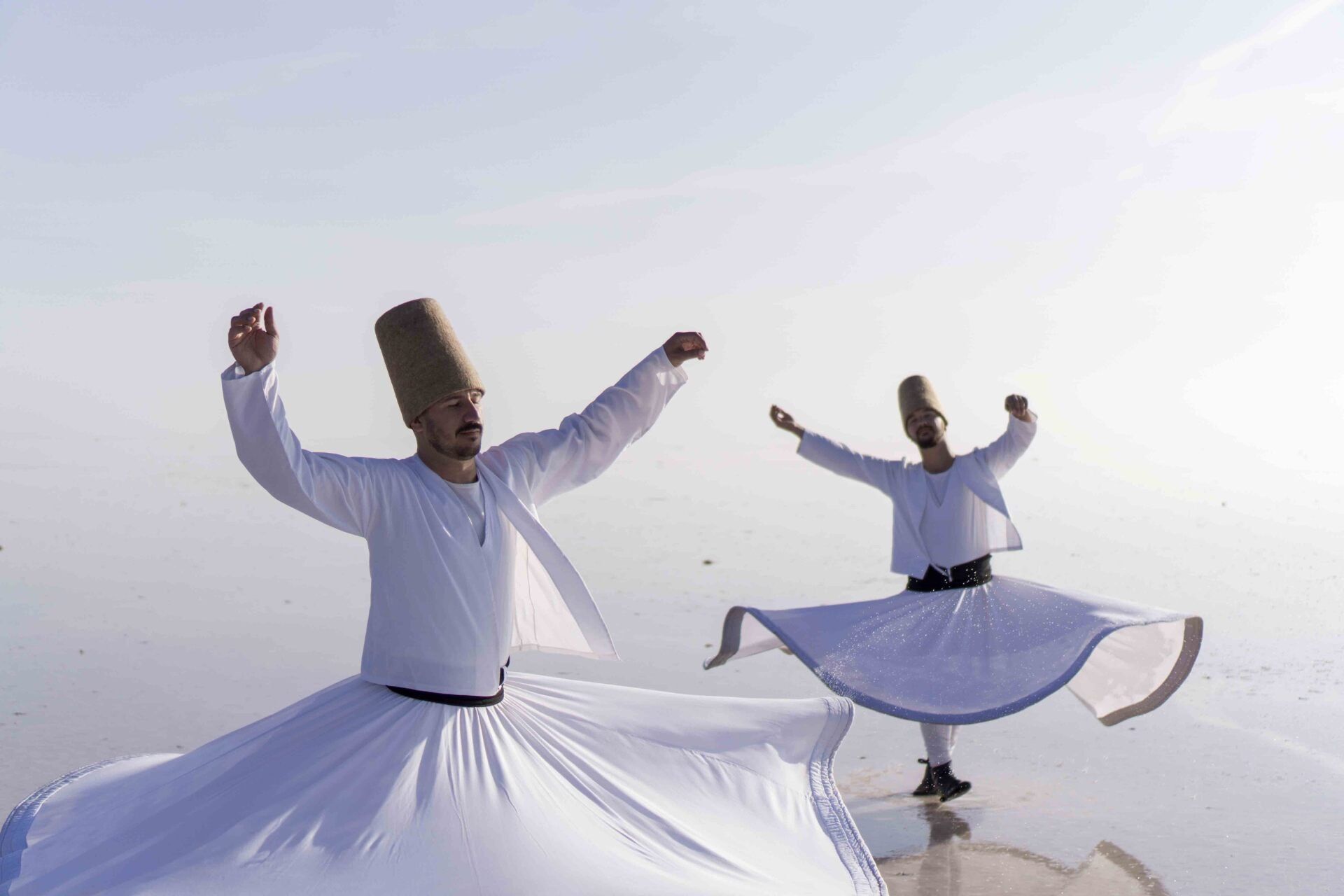 Two men in white robes are dancing in front of a body of water.