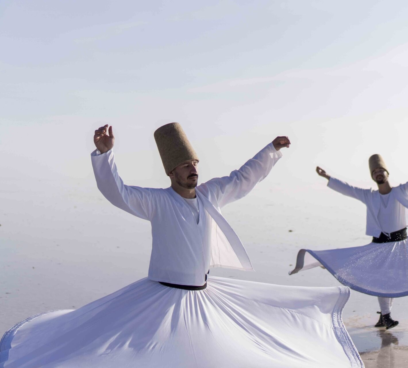 Two men in white dresses and hats are dancing on a beach.
