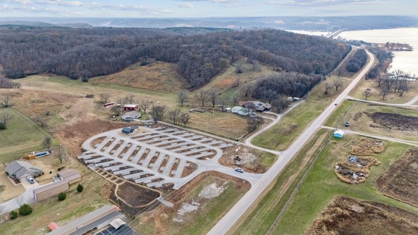 Aerial view of a paved boat ramp parking lot next to a road, field, and forested hillside near a body of water.