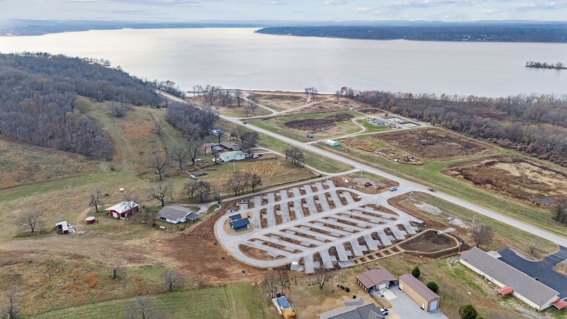 An aerial view of a rural gravel parking lot near a large body of water, surrounded by hills and fields.