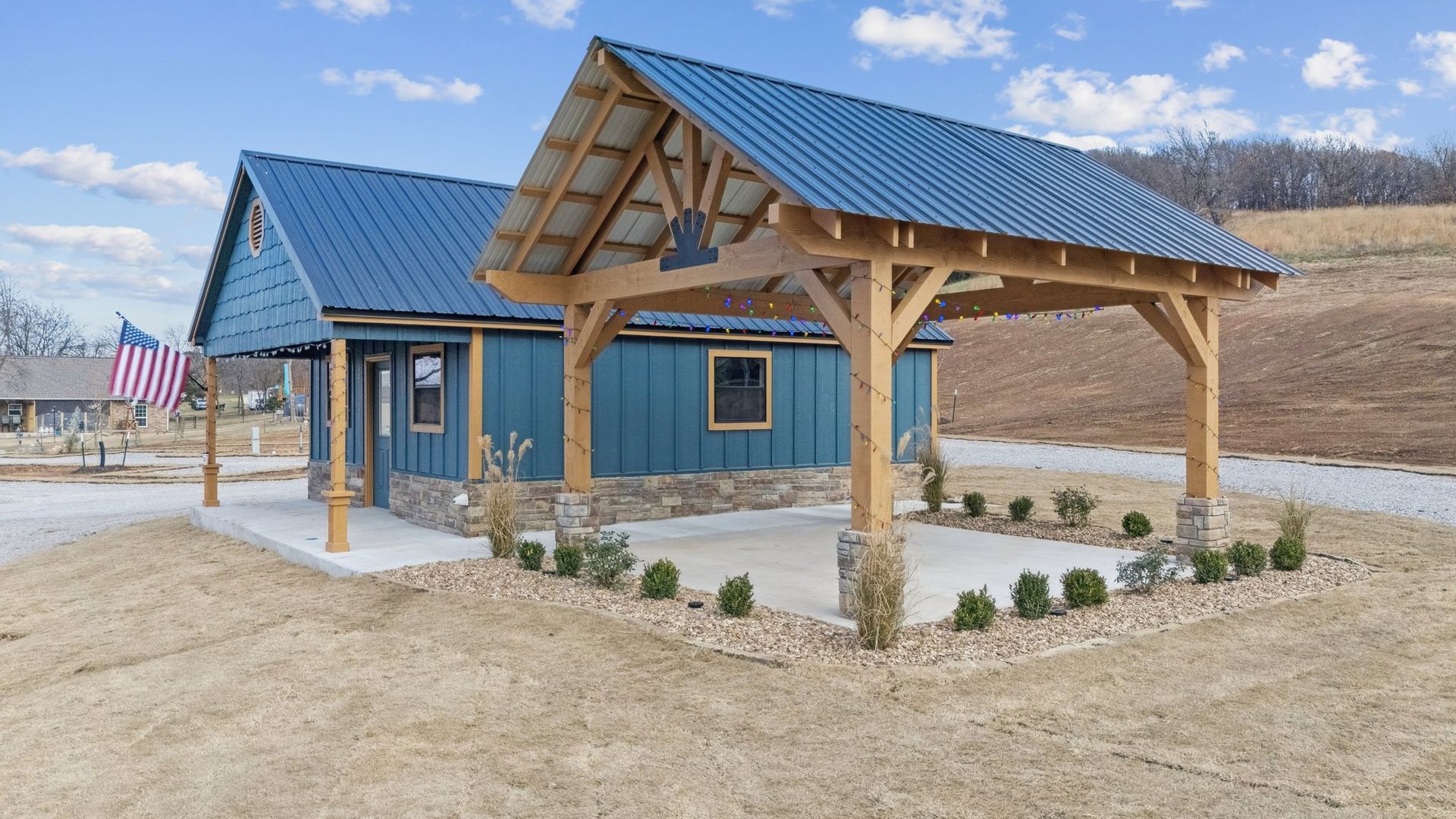 Blue-sided cabin with a metal roof and a large wooden-framed open pavilion attached to the side, set in a rural field.