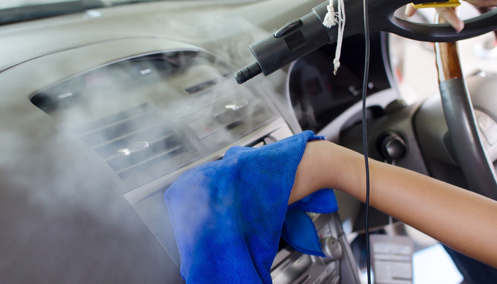 Person cleaning a car dashboard with a steam cleaner and blue cloth.