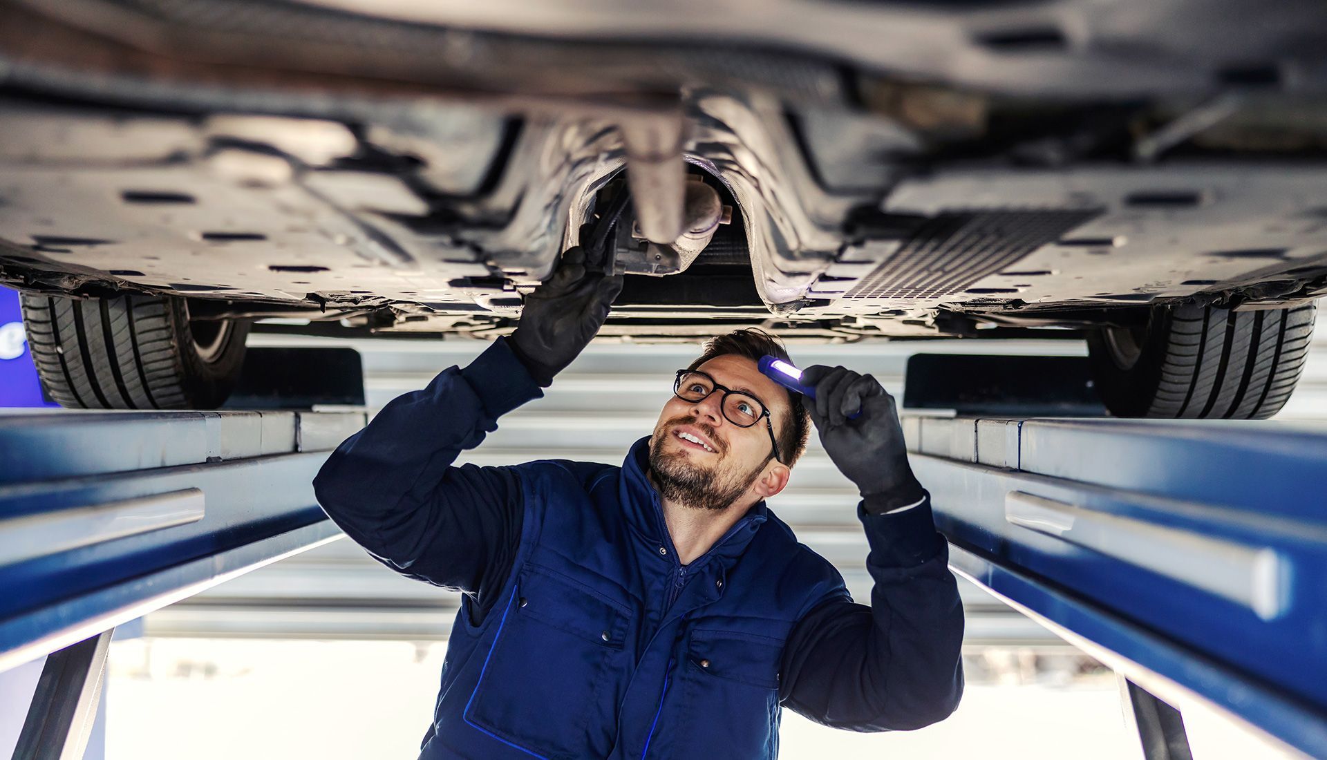 Mechanic inspecting car underside with a flashlight, smiling.