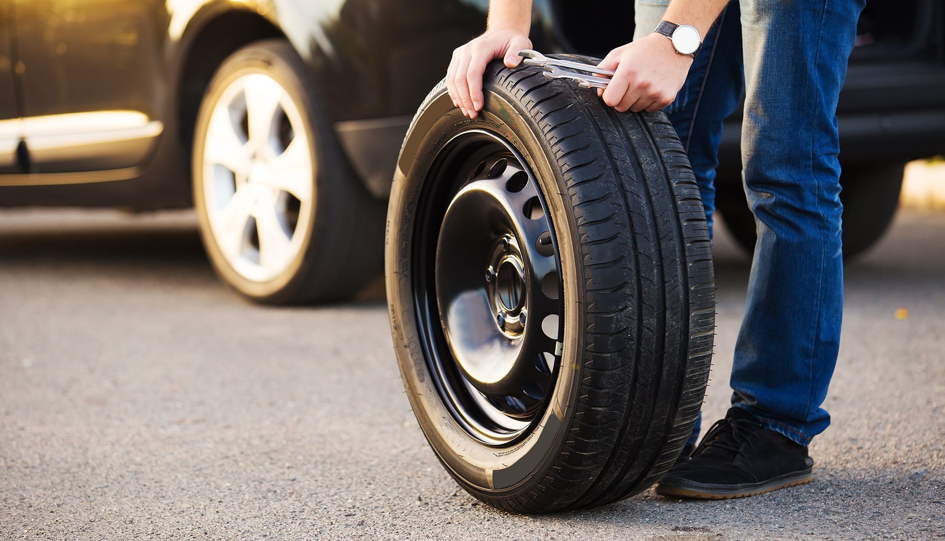 Person holding a spare tire next to a black car on a paved road.