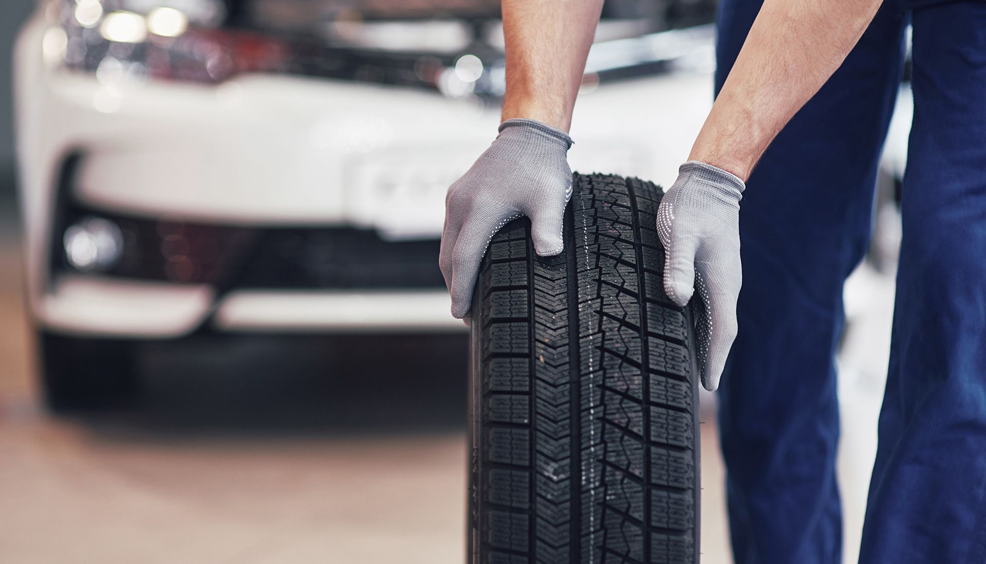 Mechanic holding a car tire in front of a white car.