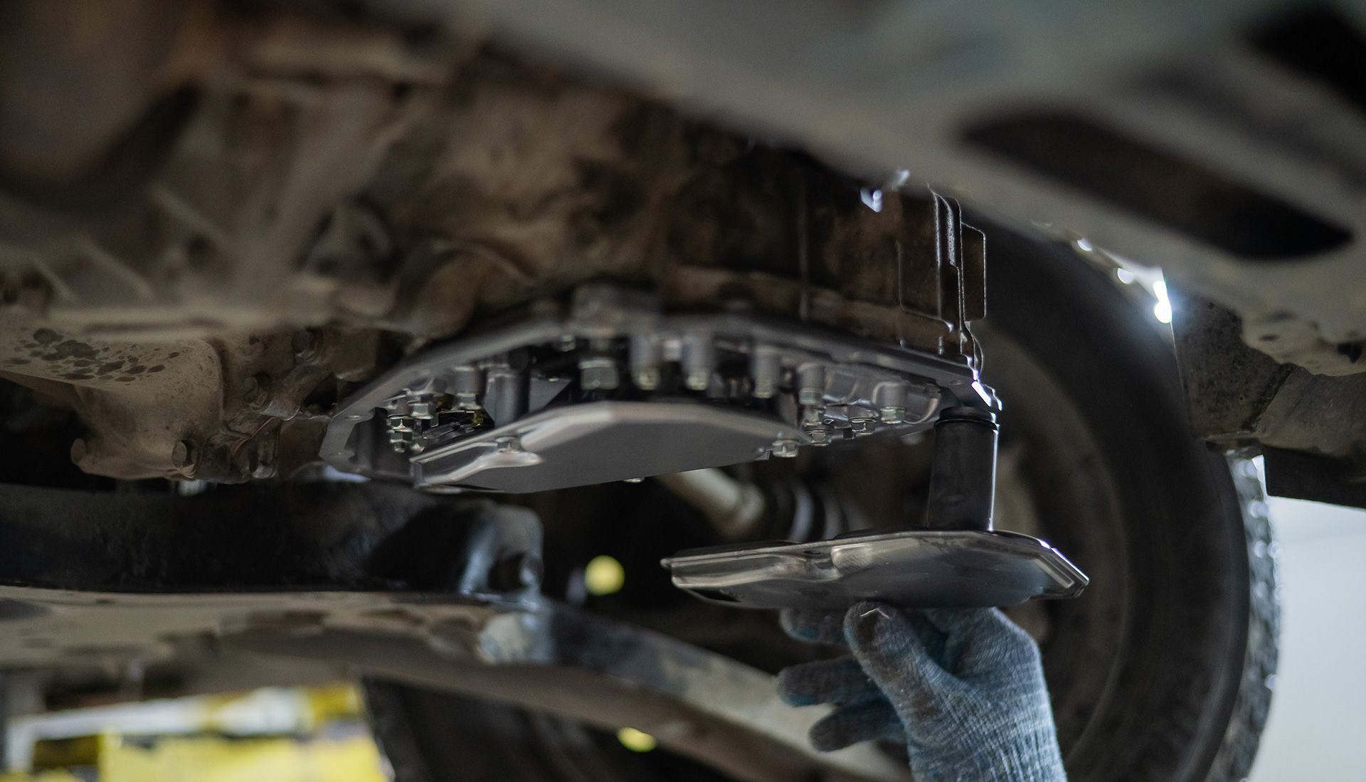 Person removing transmission pan from vehicle, dripping oil. Under vehicle, gloves visible.