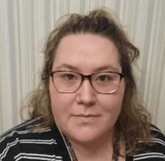 Woman with glasses and curly hair in front of a striped curtain. She is wearing a striped shirt and looking at the camera.