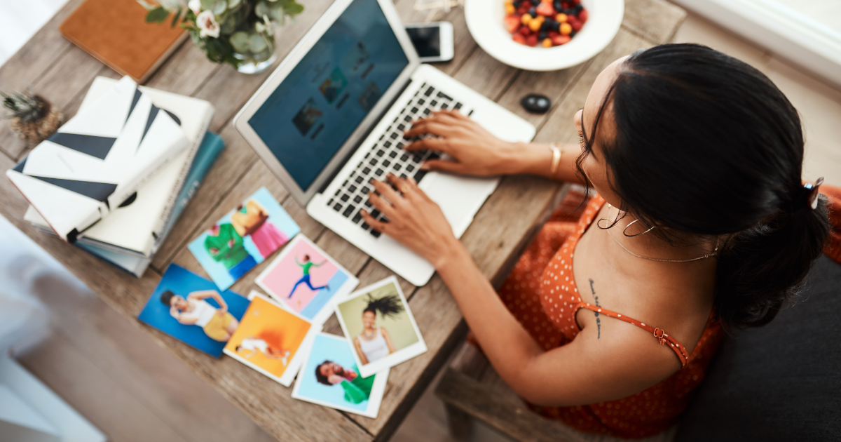 Overview of woman typing on laptop with colorful photos around surrounding the tabletop. 