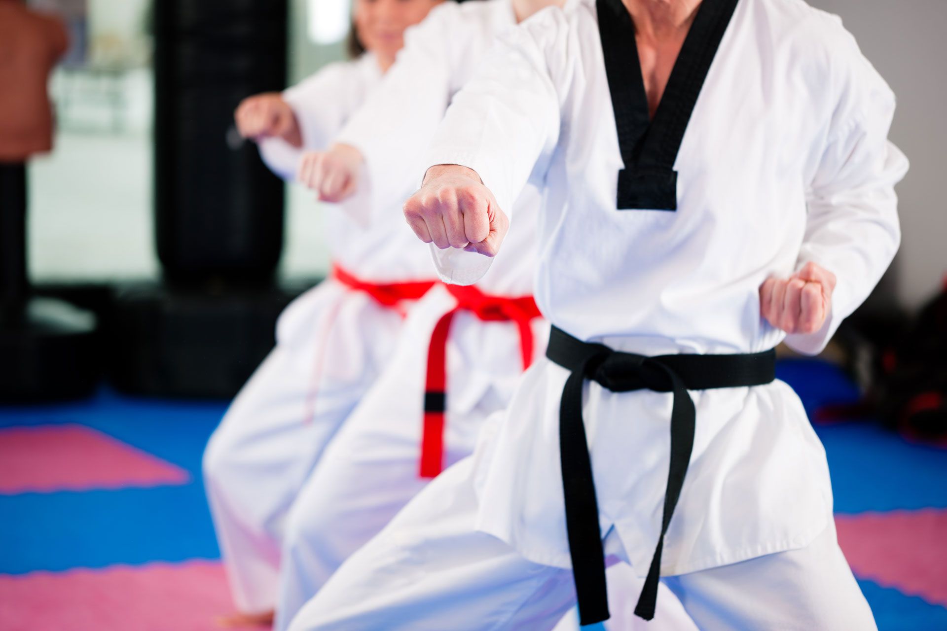 Three martial artists in white uniforms practicing punches in a studio, wearing black and red belts.
