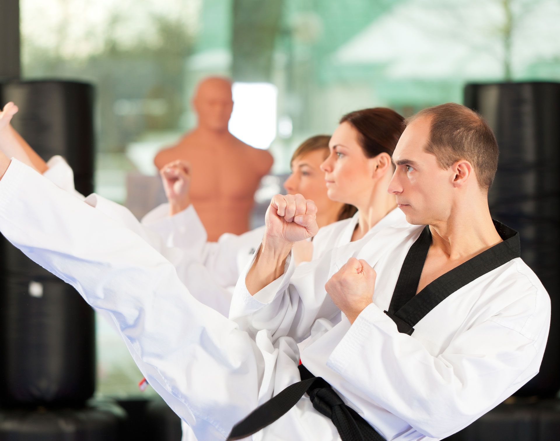 Three people in black-belt martial arts uniforms practice side kicks in a gym setting with punching bags.