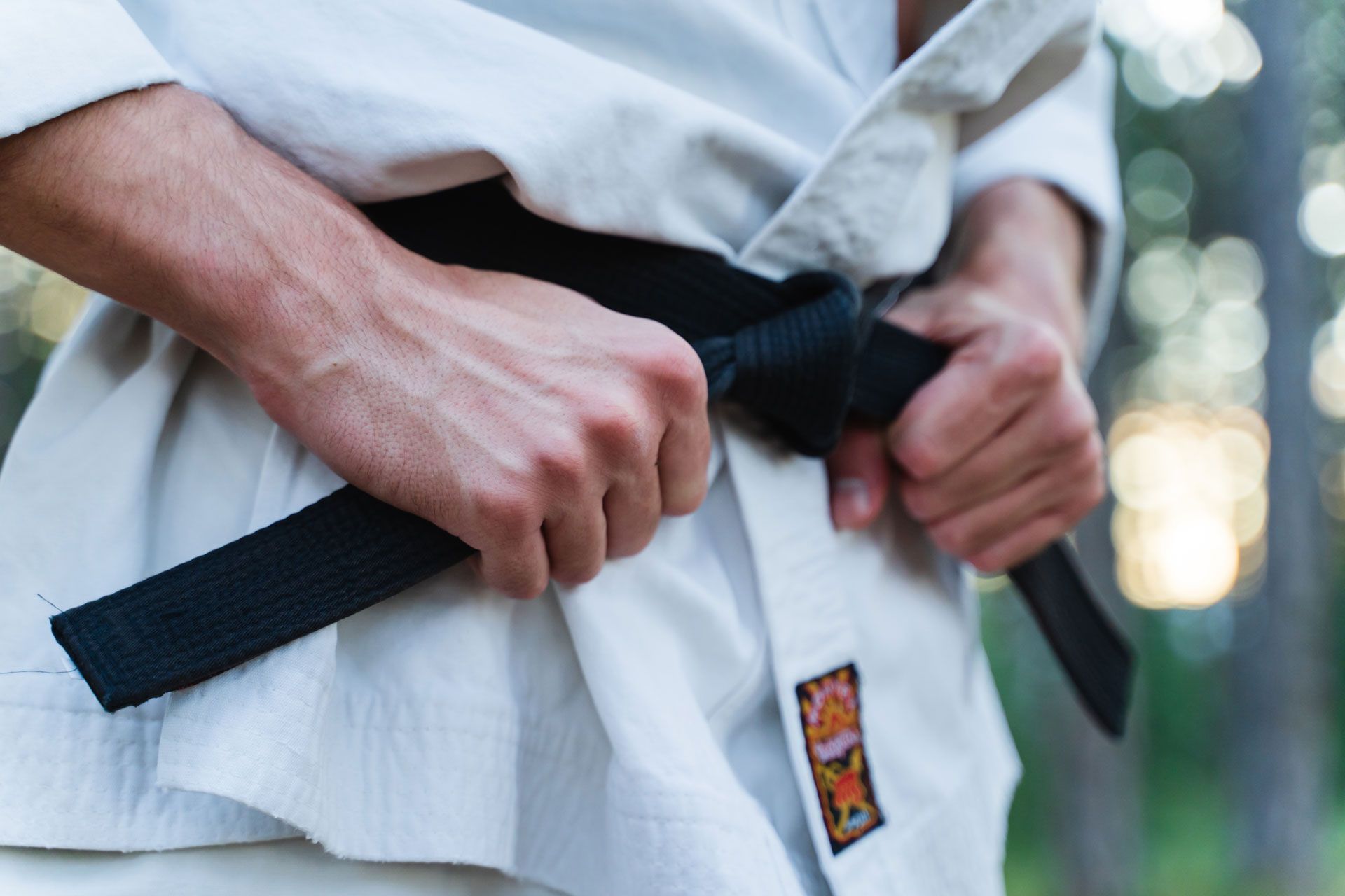 Close-up of a person in a white martial arts uniform adjusting a black belt outdoors with a blurred, natural background.