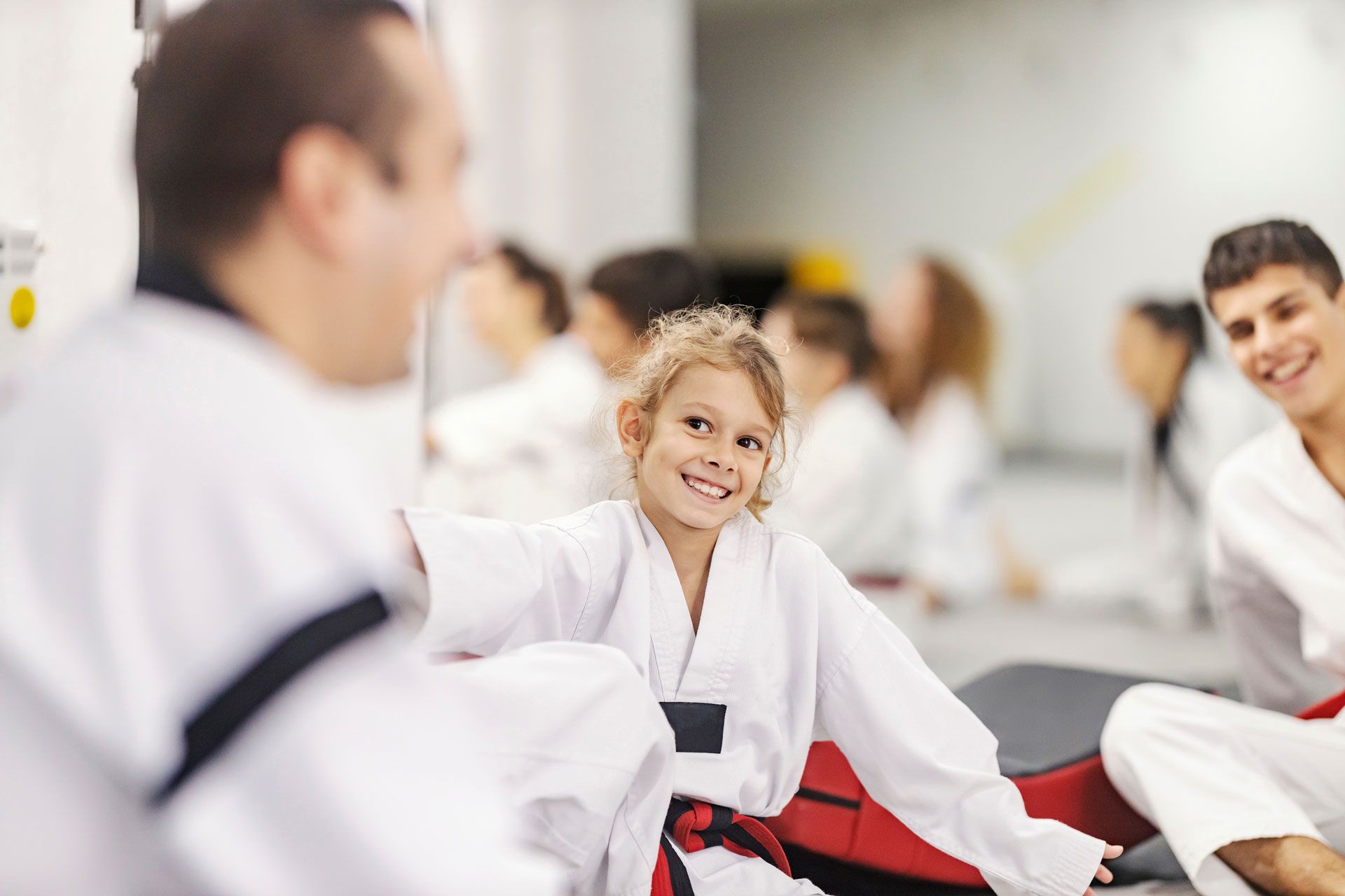 A smiling person in a white martial arts uniform sits in a training hall among others wearing similar gear.