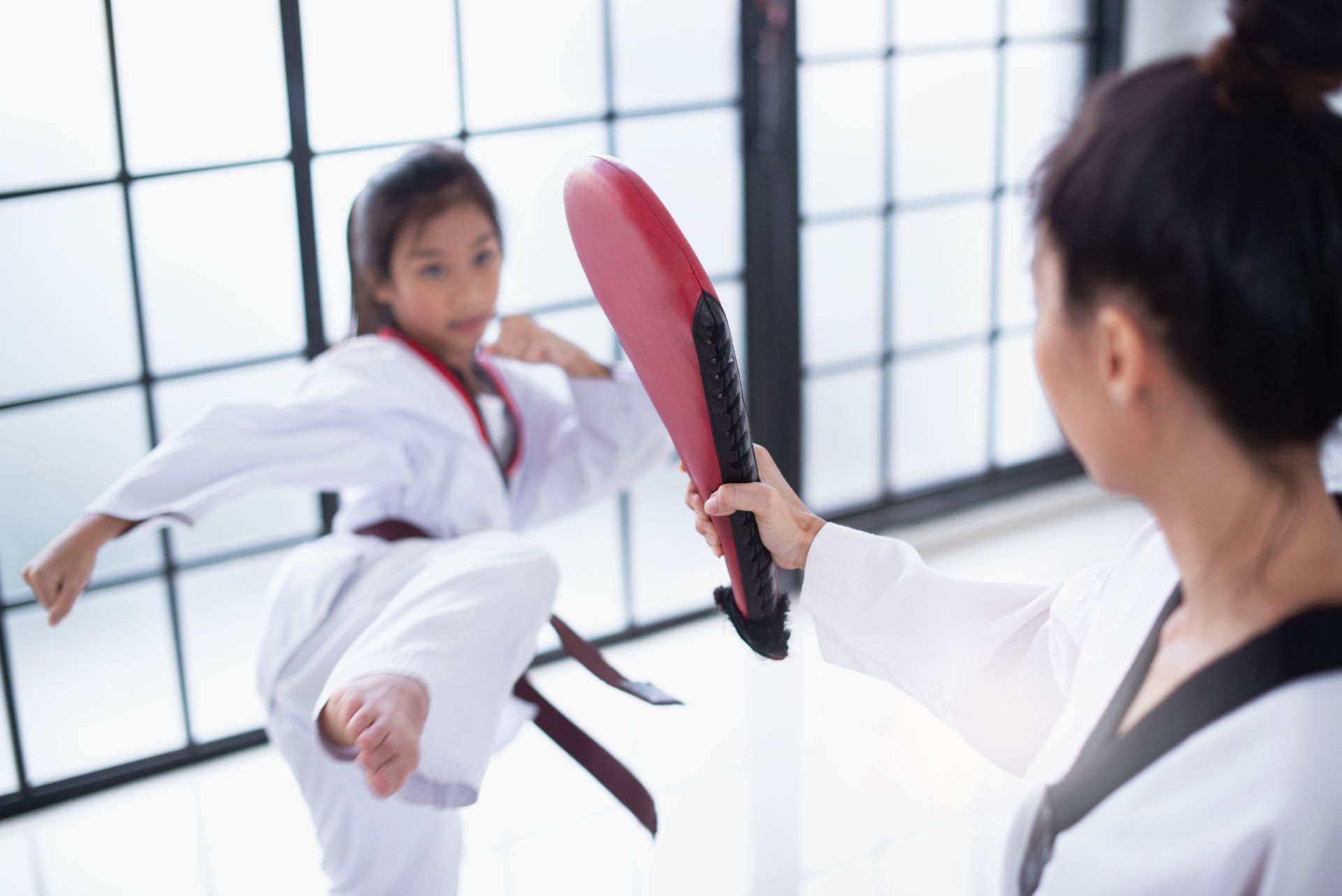 A person in a martial arts uniform kicks a red training paddle held by an instructor in a bright studio.