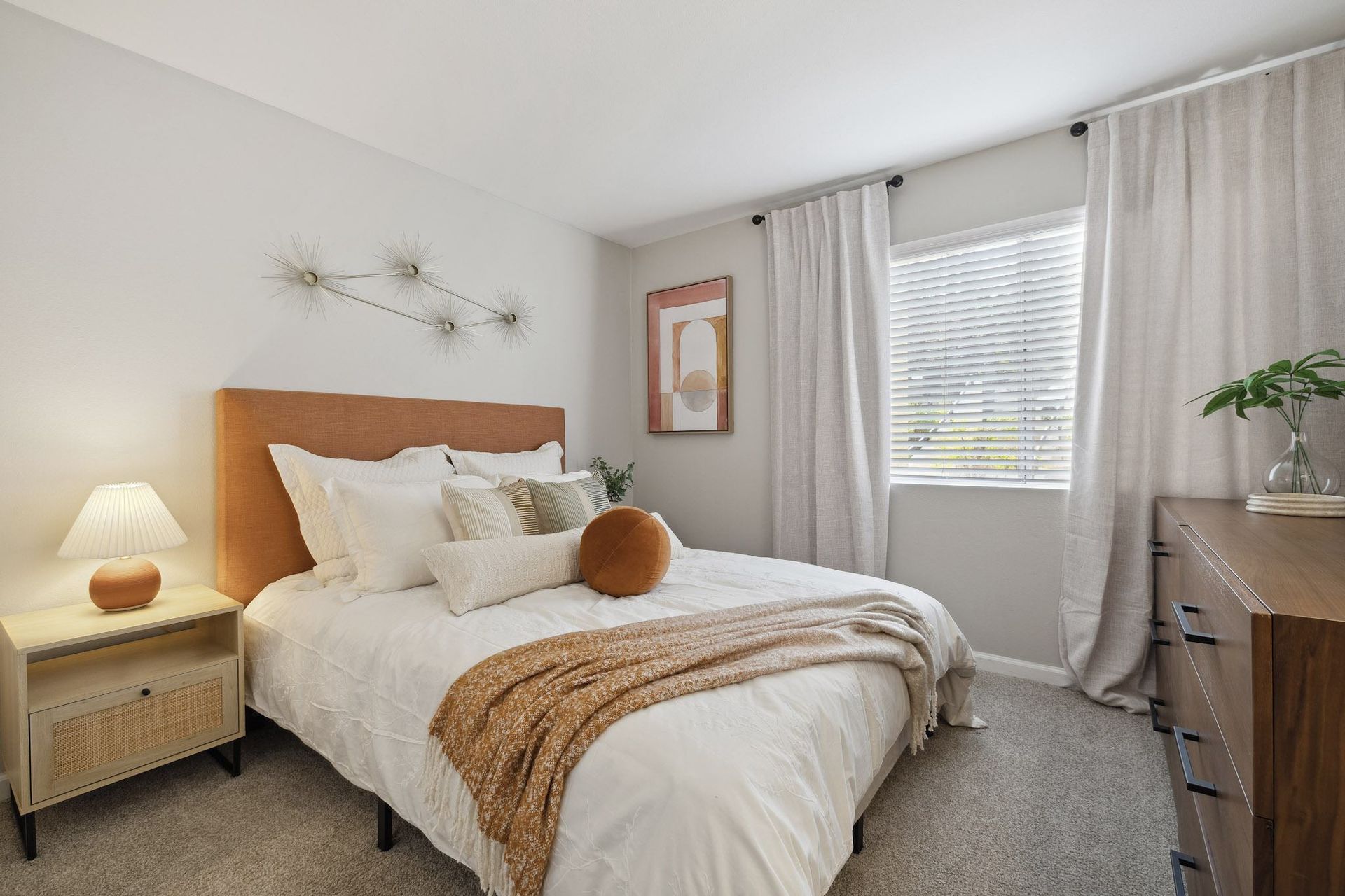 Bedroom in a rental unit featuring a white bed, tan headboard, nightstand lamp, dresser, and window with blinds.