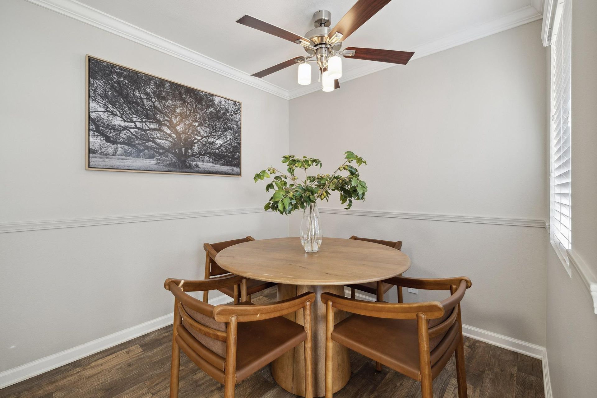 Dining nook with round wooden table, six chairs, ceiling fan, and framed tree artwork.