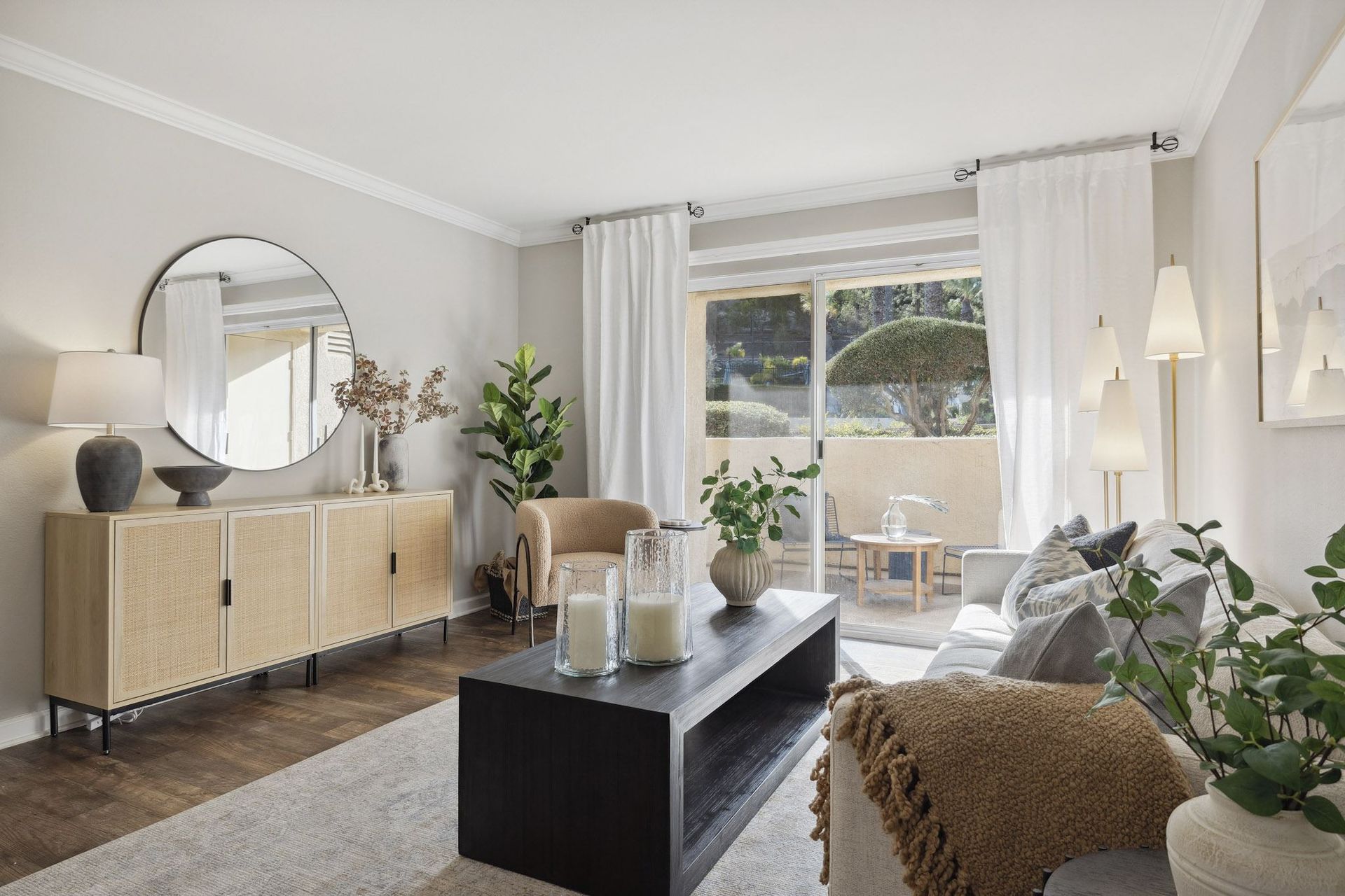 Living room in an apartment with sliding glass doors, a round mirror, neutral furniture, and plants.