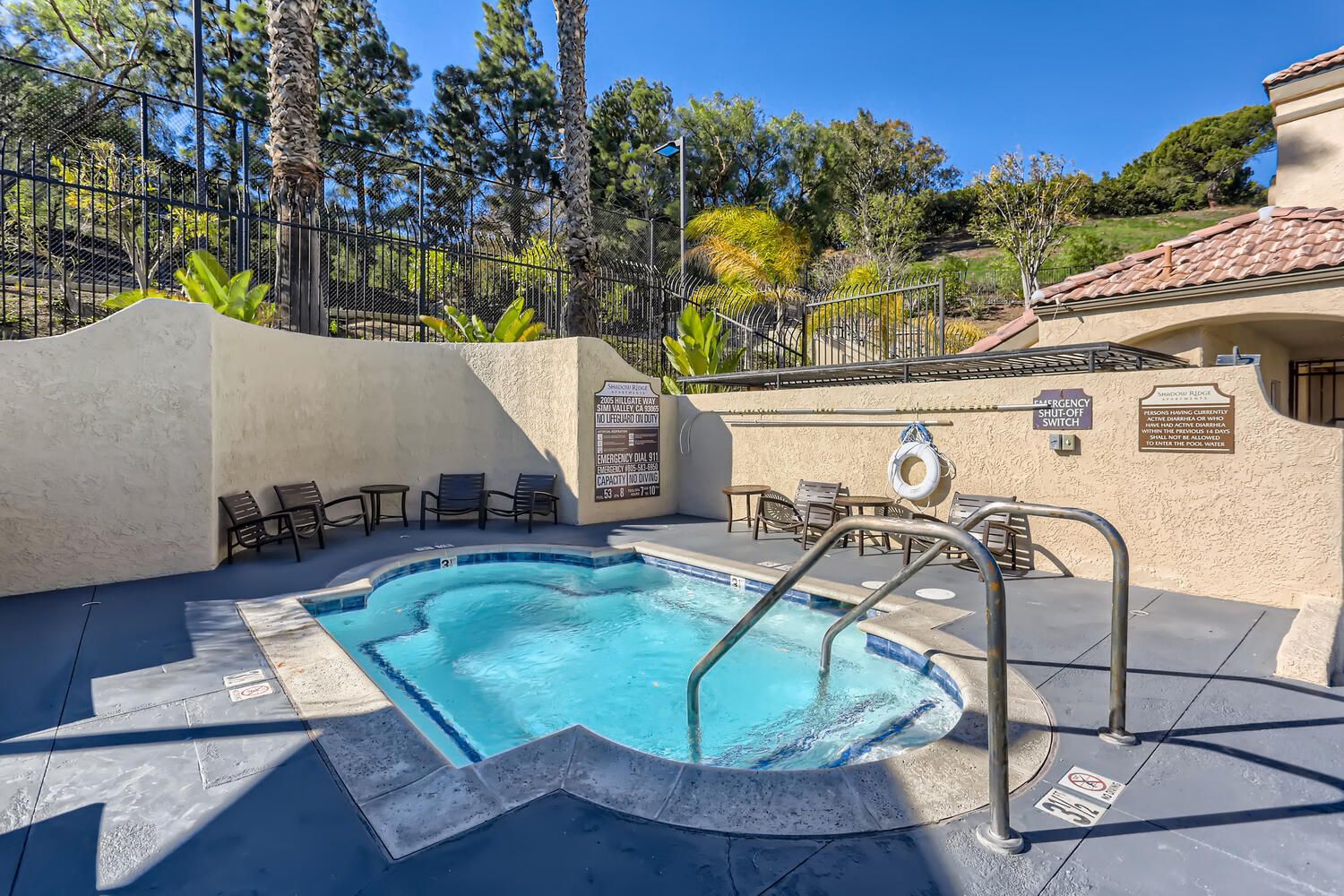 Outdoor community pool area with curved blue pool, lounge chairs, and a tall fence.