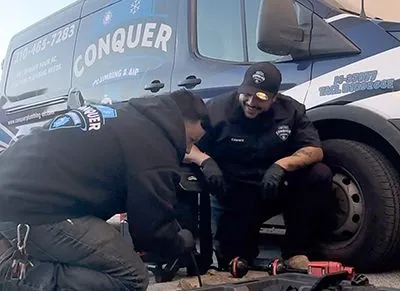 Two men are working on a car in front of a conquer van.