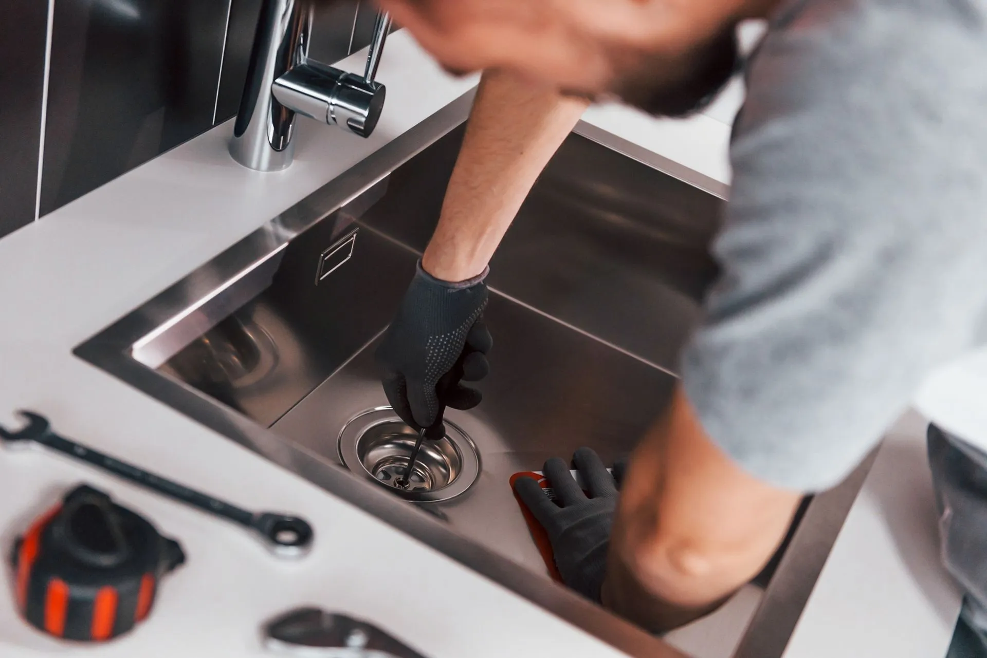 Plumber working on a kitchen sink, wearing black gloves, tools nearby.