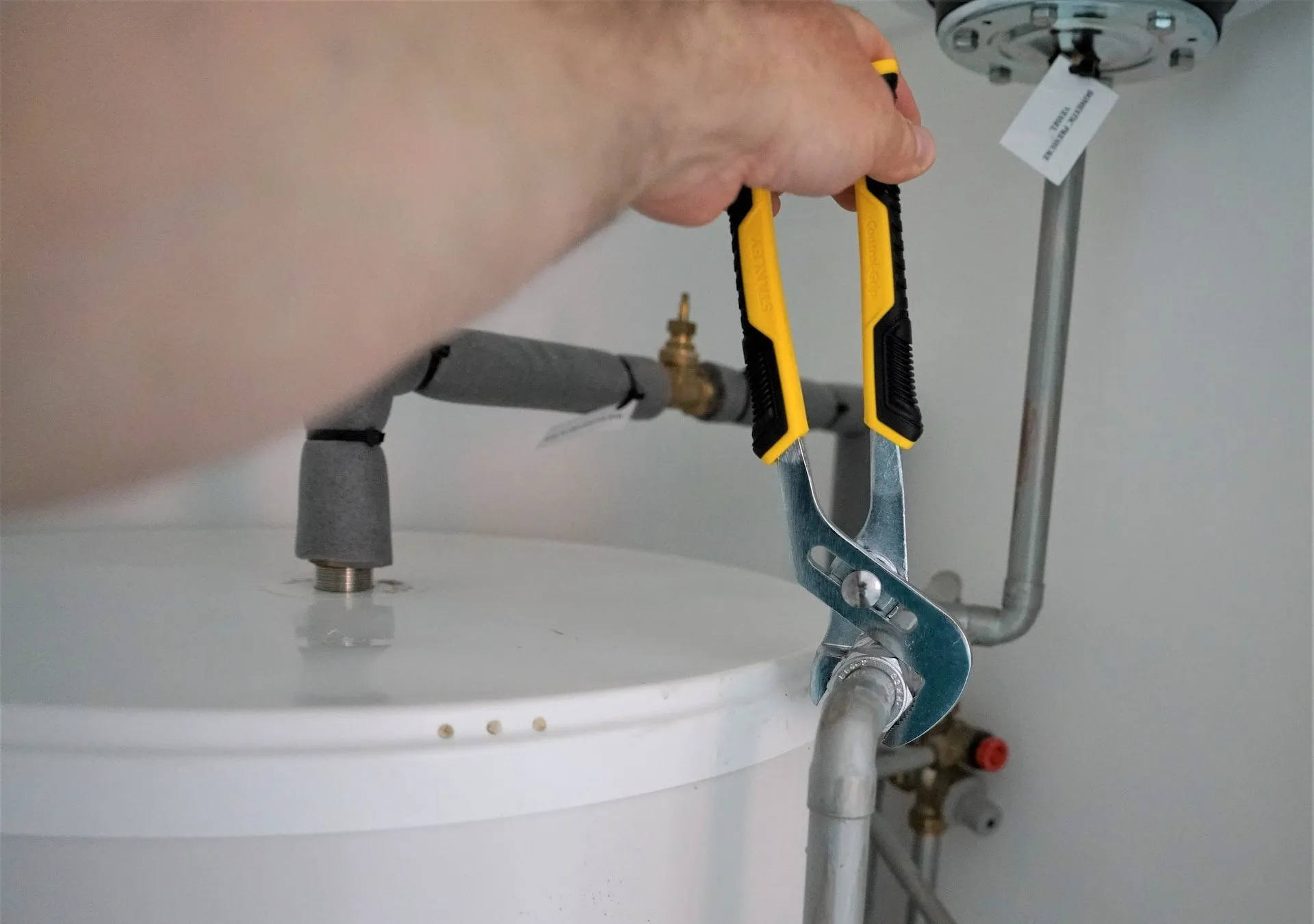 A person uses yellow-handled tongue-and-groove pliers to tighten a metal pipe fitting on a white water heater.