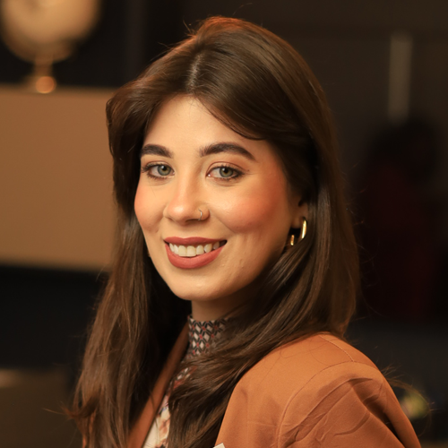 Uma mulher vestindo uma jaqueta marrom e brincos está sorrindo para a câmera.