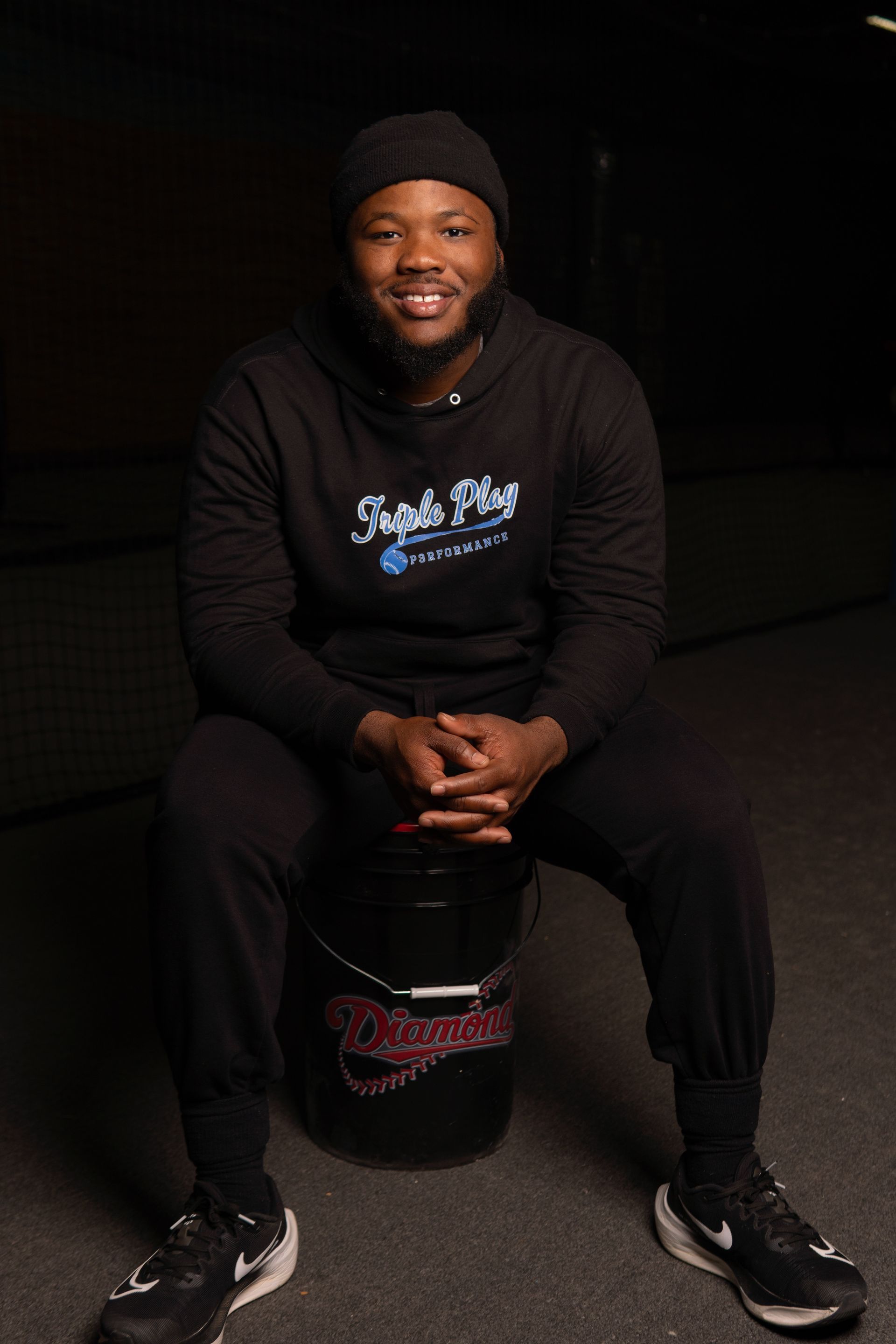 A man is sitting on a black barrel with his hands folded.