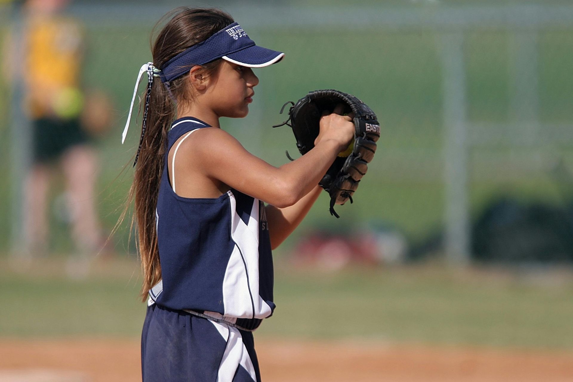 A little girl is wearing a visor and holding a baseball glove.