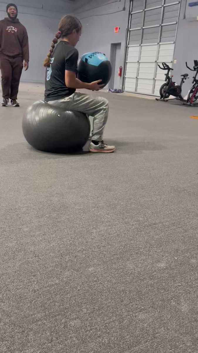 A woman is sitting on an exercise ball in a gym holding a ball.