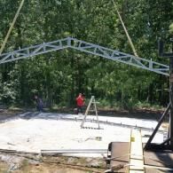 Workers assembling a metal truss frame on a concrete slab in a wooded outdoor area