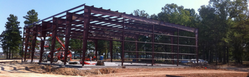 Steel frame building under construction on a dirt site with trees in the background
