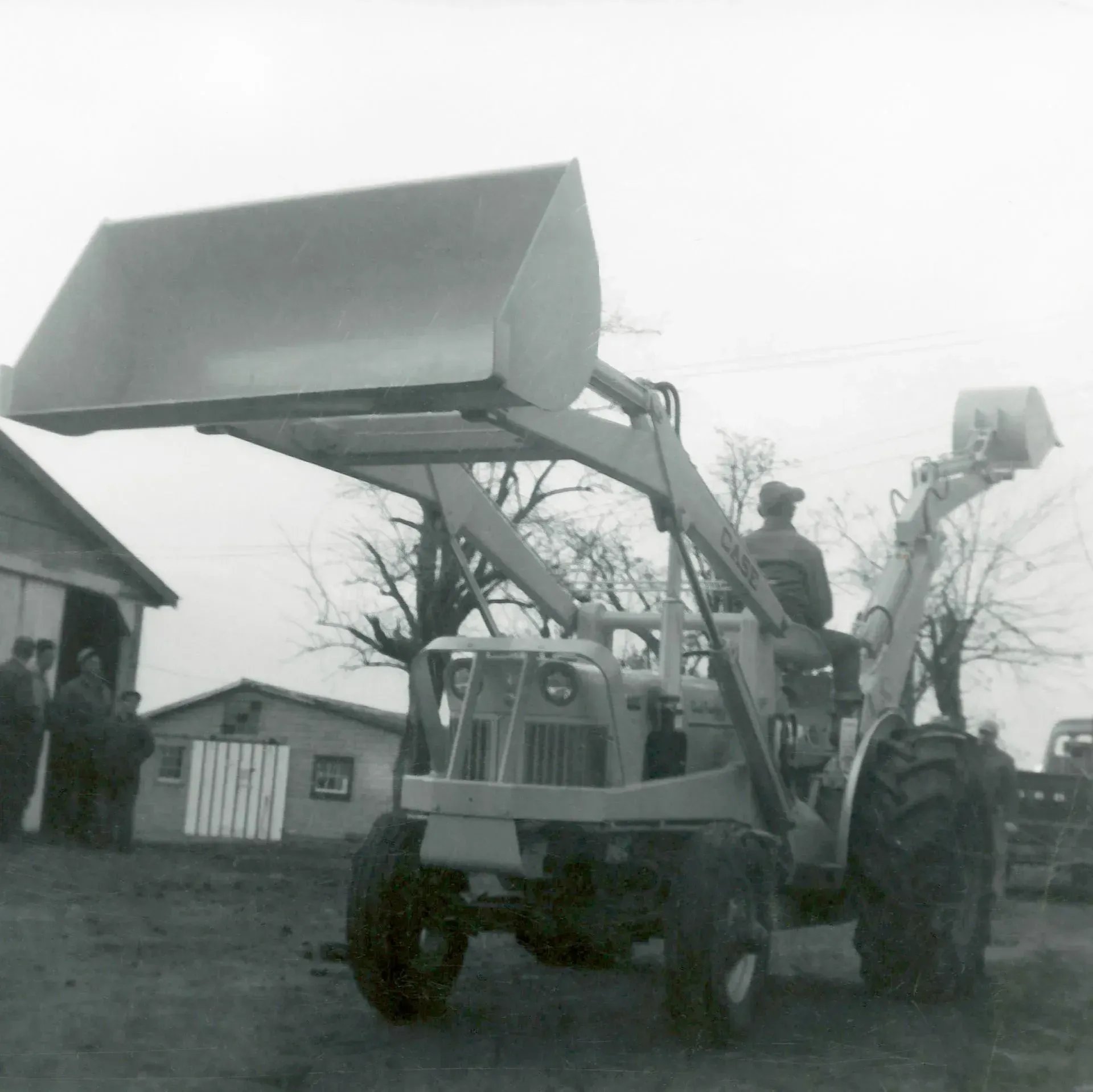 A light-colored tractor with a raised bucket, driven by a person, in front of a barn.