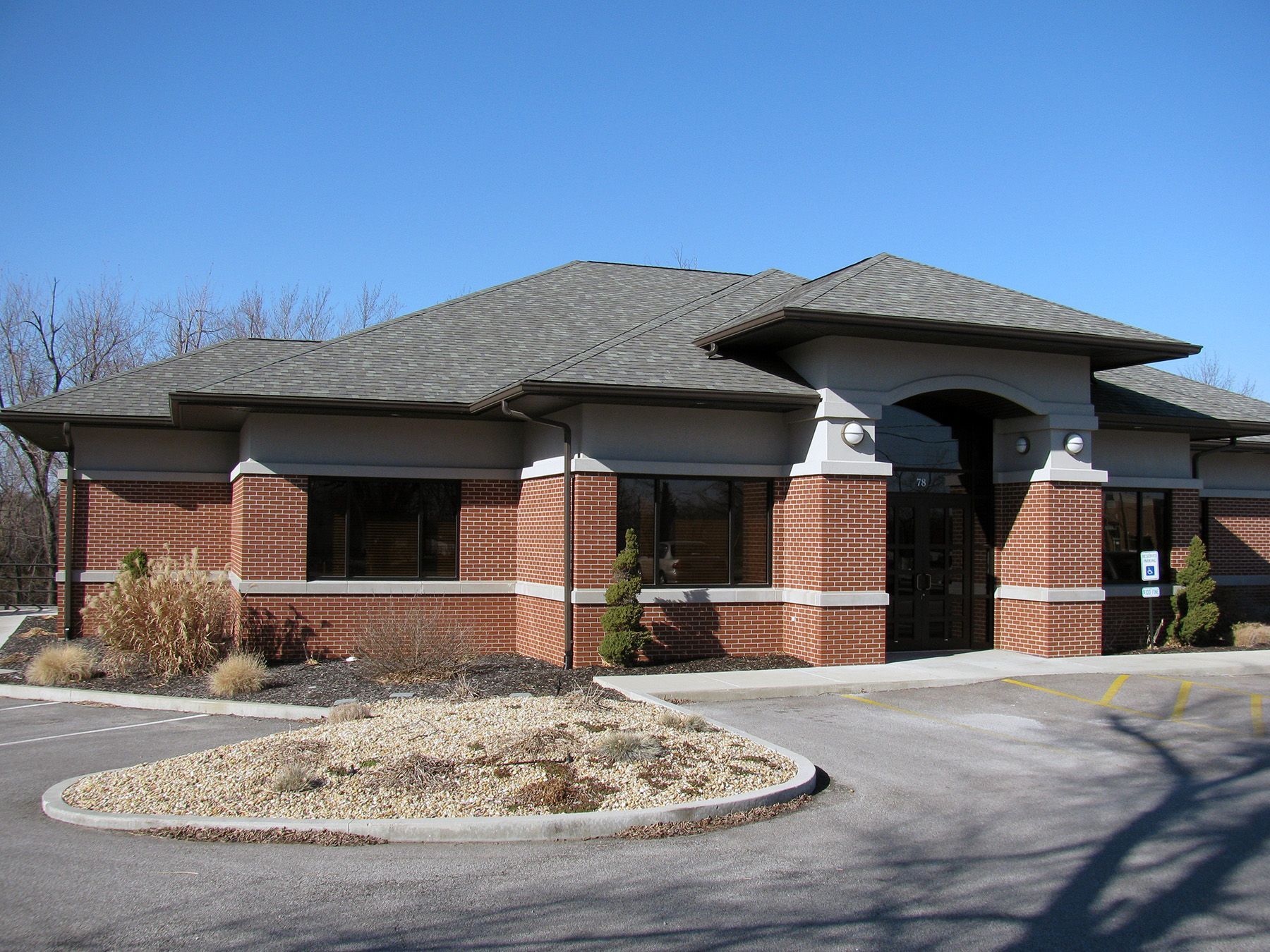 Brick building with dark windows and gray roof; small landscaped island in front.