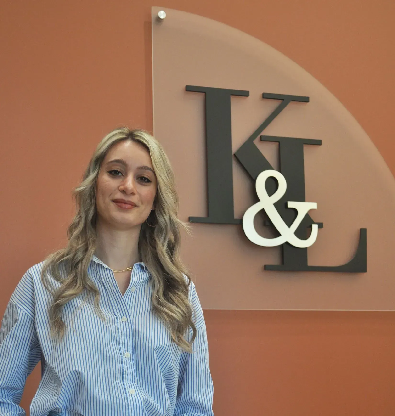 Woman with blonde hair wearing a striped shirt stands by a wall with a logo that says