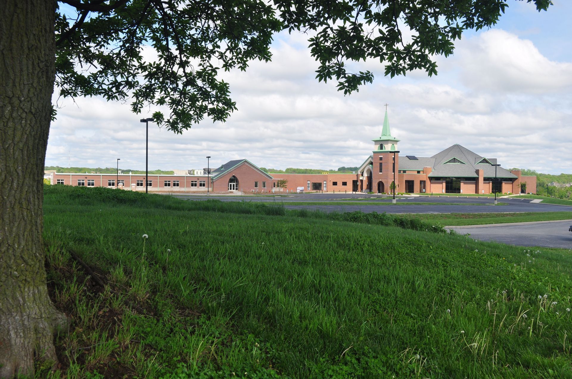 Brick building with a green steeple, behind a grassy hill and tree. Cloudy sky.