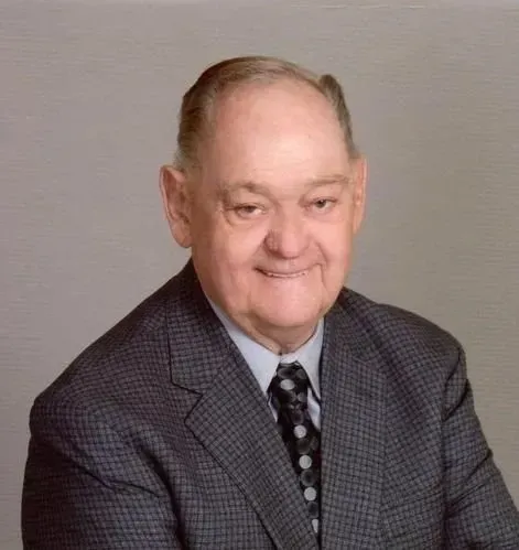 Elderly man in a suit smiles, dark tie, short hair. Studio shot with a light gray background.