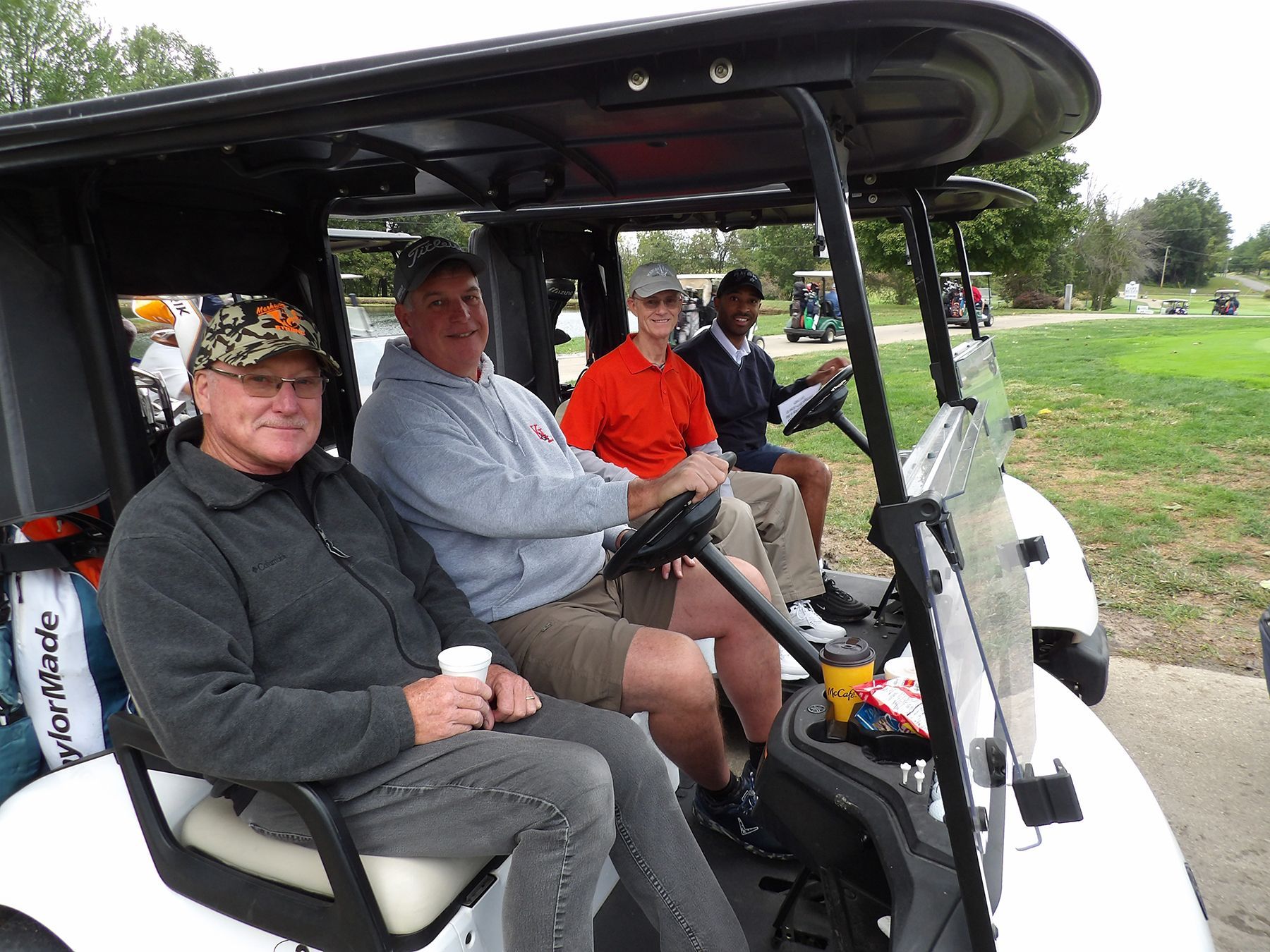 Four men in golf attire pose by golf carts. Two men wear red shirts, two wear black and white, all smile.