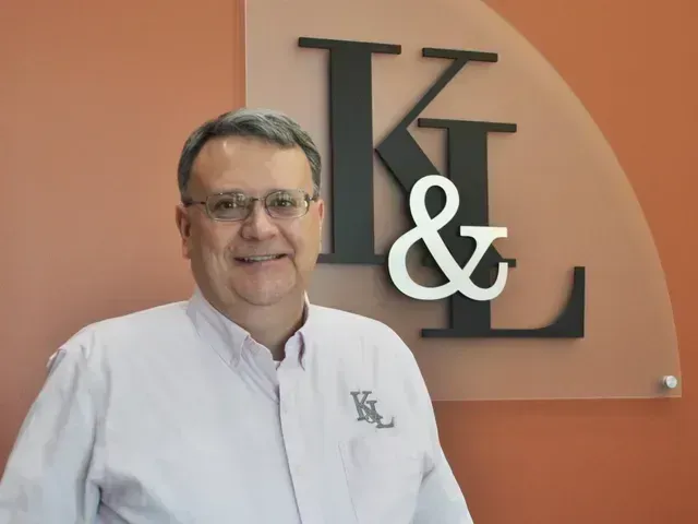 Man in glasses and light pink shirt smiles in front of a wall with
