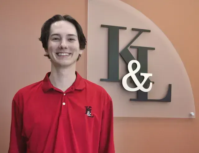 Young man smiles in red shirt with logo, standing in front of the