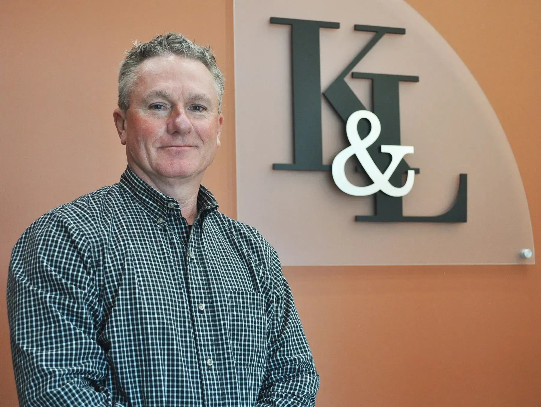 Man in checked shirt smiles, standing beside a wall with