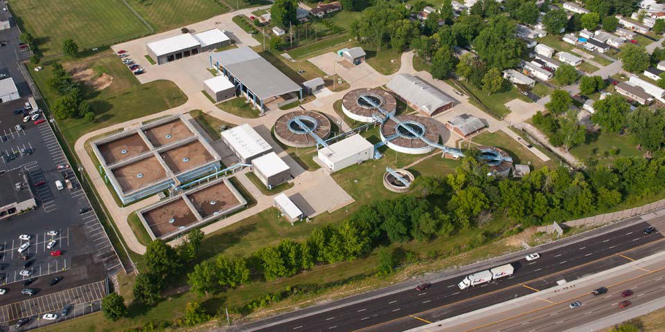 Aerial view of a wastewater treatment plant with several tanks and buildings, next to a highway.