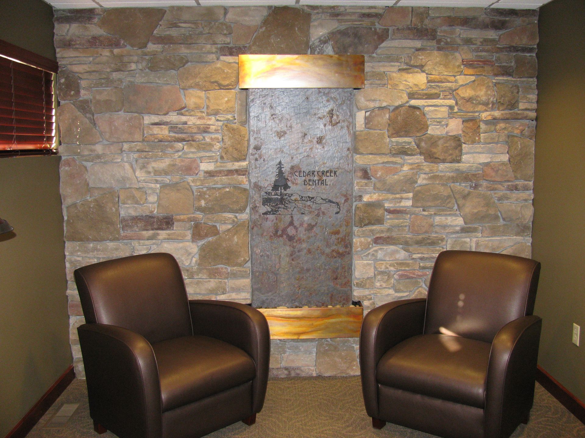 Two brown leather chairs face a stone wall with a water feature, in a room with brown shades.