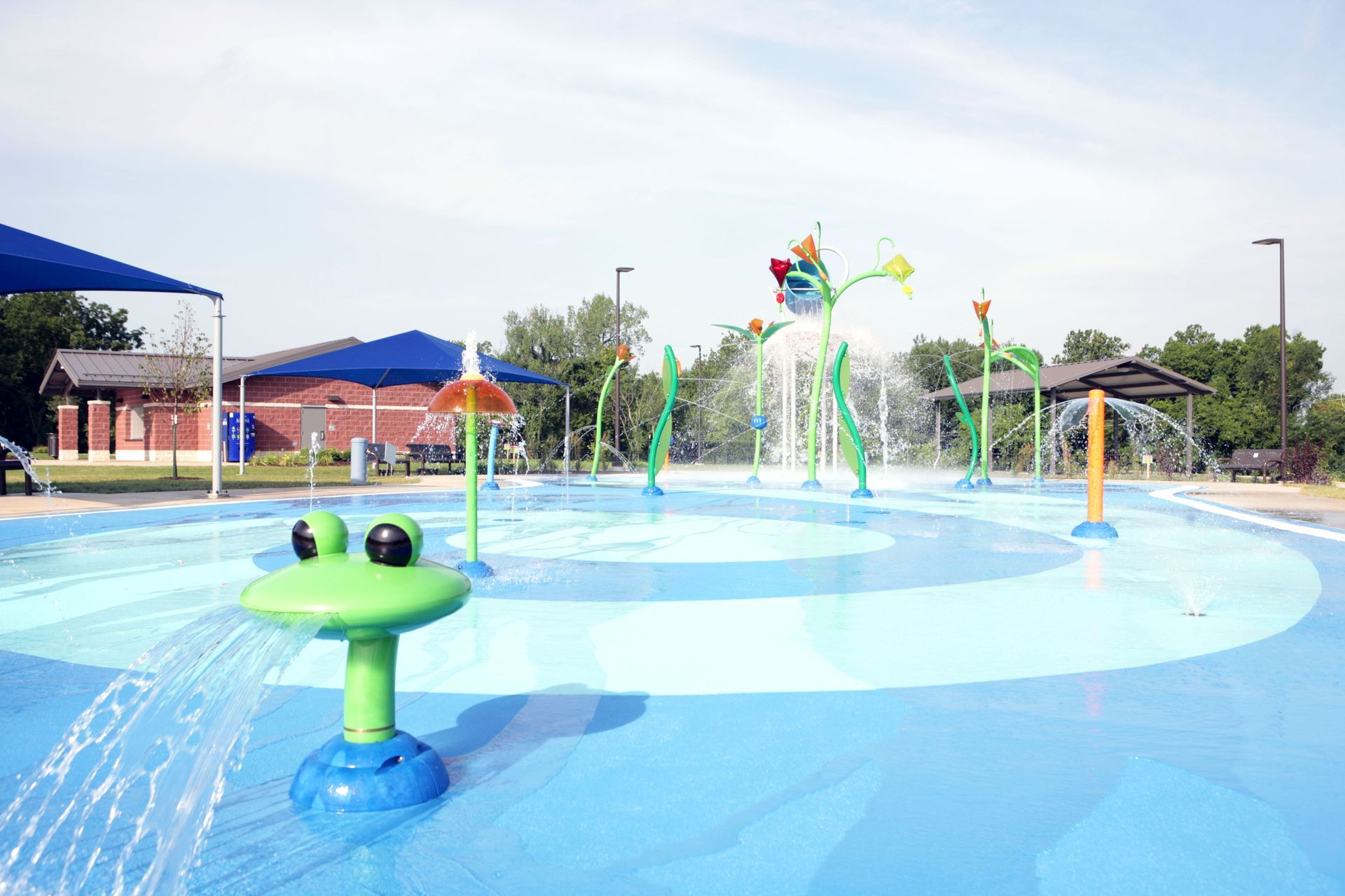 A splash pad with water features, a blue surface, and a green frog-shaped fountain under a sunny sky.