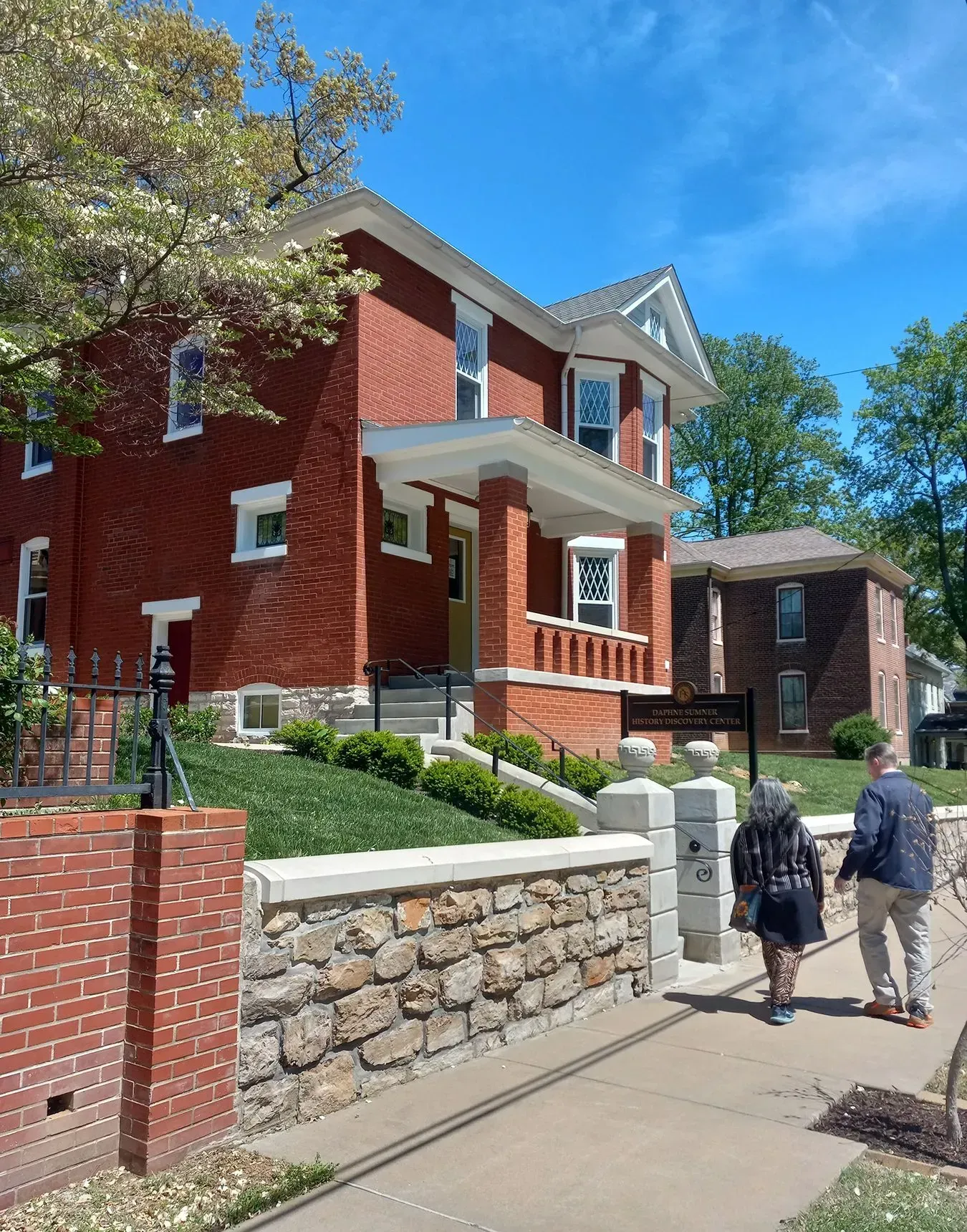Red brick house with stone wall, people walking. Sunny day.