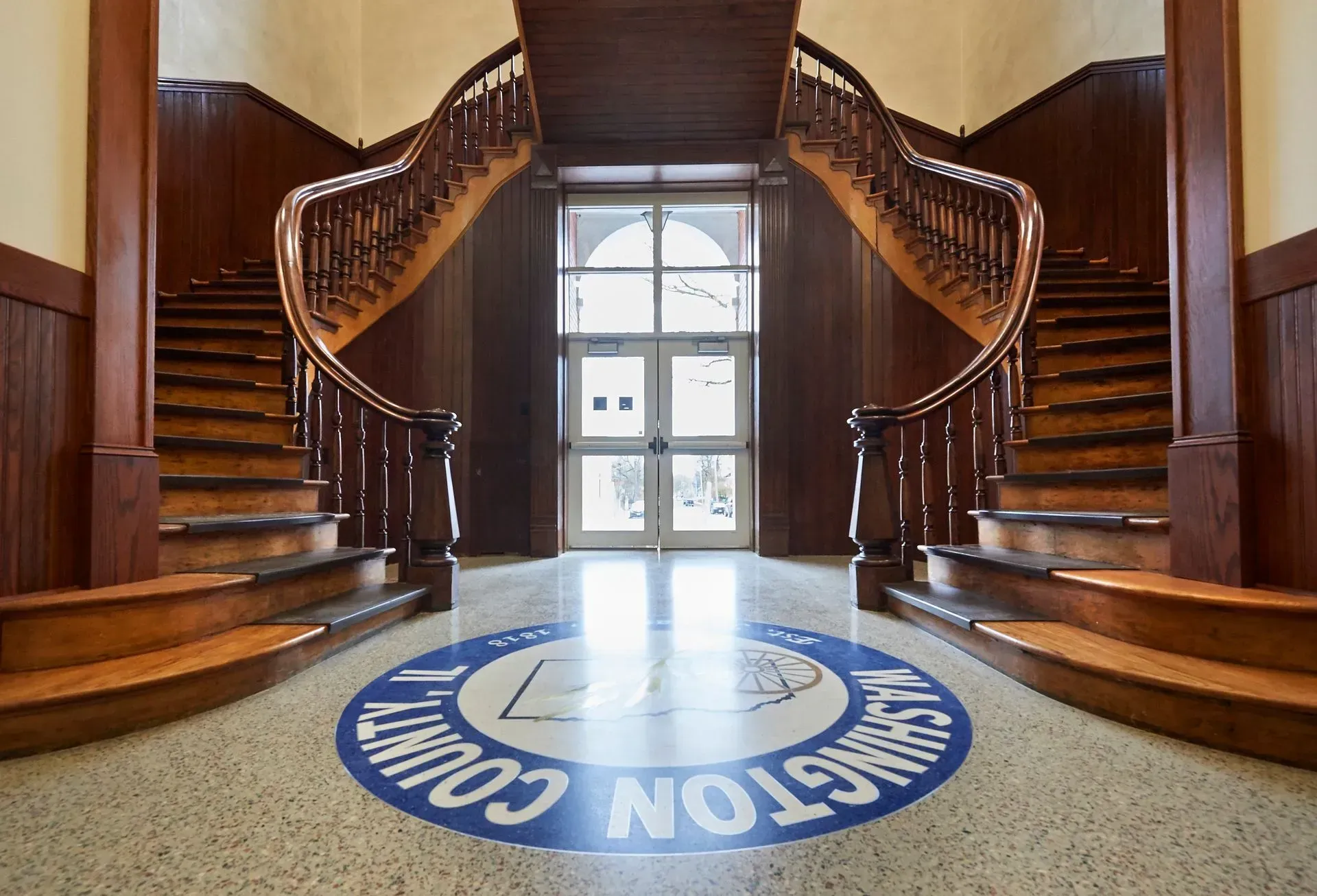 Interior with two wooden staircases flanking a doorway. A blue logo is on the floor.